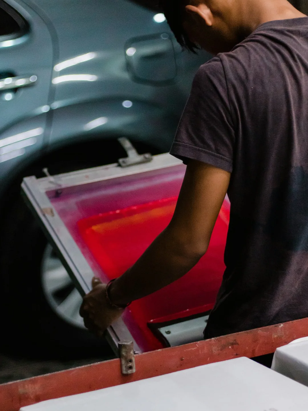 Manos levantando un marco de serigrafía cargado de tinta roja sobre la mesa de impresión